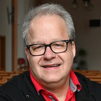 Ein lächelnder Mann mittleren Alters mit grauem Haar und Brille, der ein rotes Poloshirt und einen schwarzen Hoodie trägt.