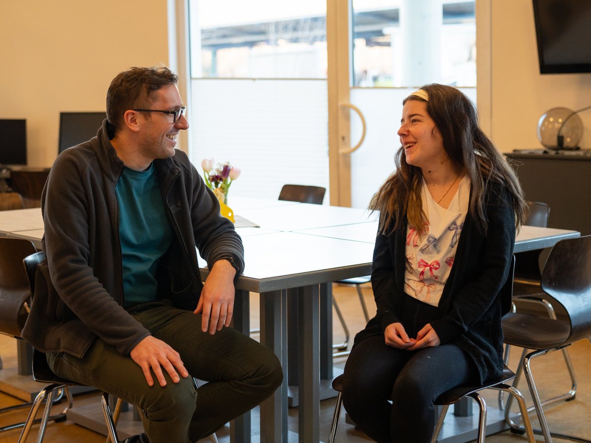 Mann und Frau lächeln und unterhalten sich an einem Tisch in einem hellen Raum.