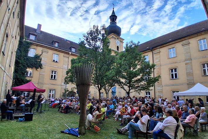 Freiluftkonzert in einem historischen Hof mit einer Band, vielen sitzenden Zuschauern auf dem Rasen und alten Gebäuden mit Turm.