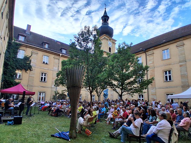 Freiluftkonzert in einem historischen Hof mit einer Band, vielen sitzenden Zuschauern auf dem Rasen und alten Gebäuden mit Turm.