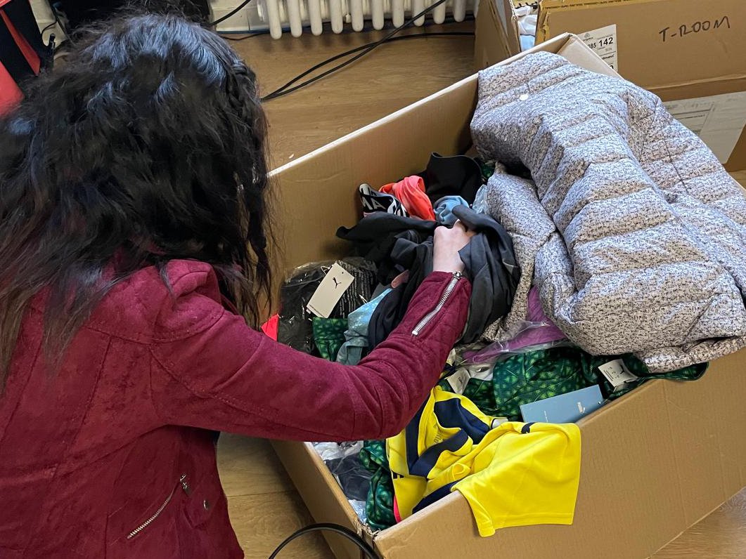 Person in roter Jacke sortiert Kleidung aus einem großen Karton, gefüllt mit verschiedenen Kleidungsstücken.