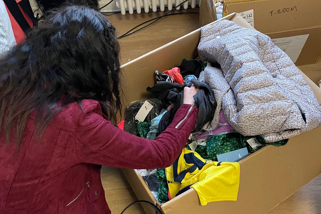 Person in roter Jacke sortiert Kleidung aus einem großen Karton, gefüllt mit verschiedenen Kleidungsstücken.