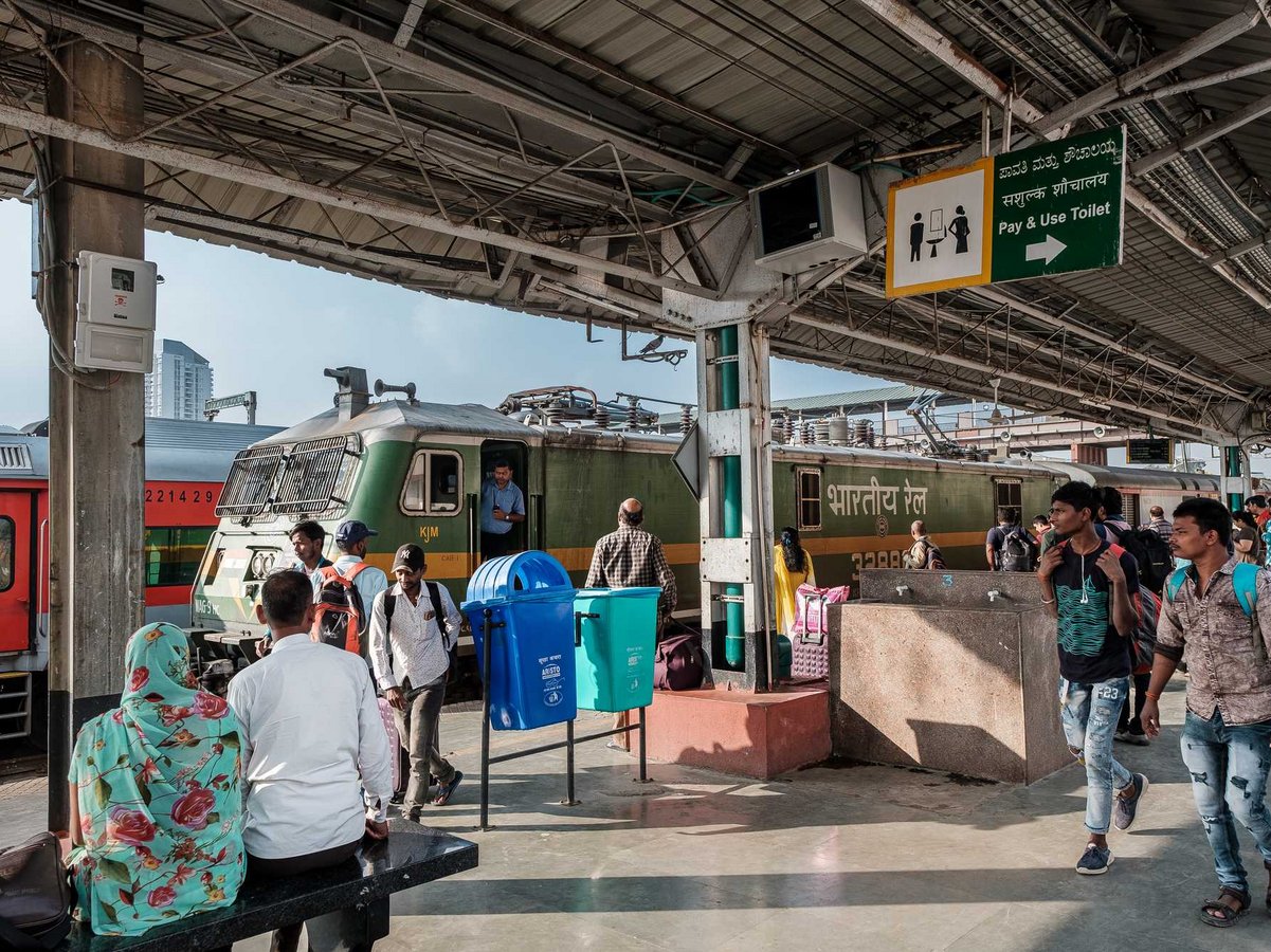 Belebter indischer Bahnsteig mit grüner Lok, roten Waggons und vielen Menschen, die gehen und warten.
