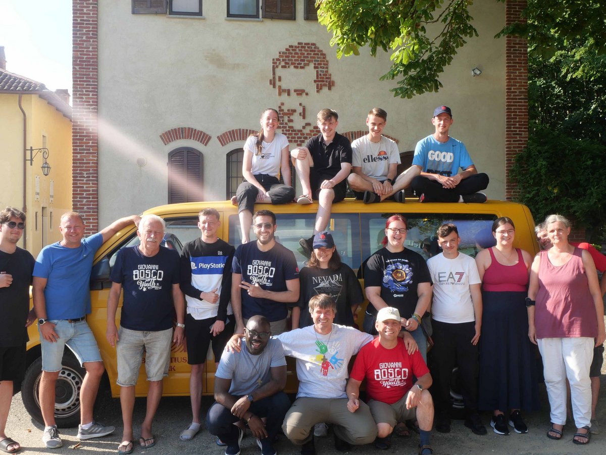 Eine lachende Gruppe von 20 jungen und älteren Menschen posiert um einen gelben Van vor einem Gebäude mit grünen Bäumen.