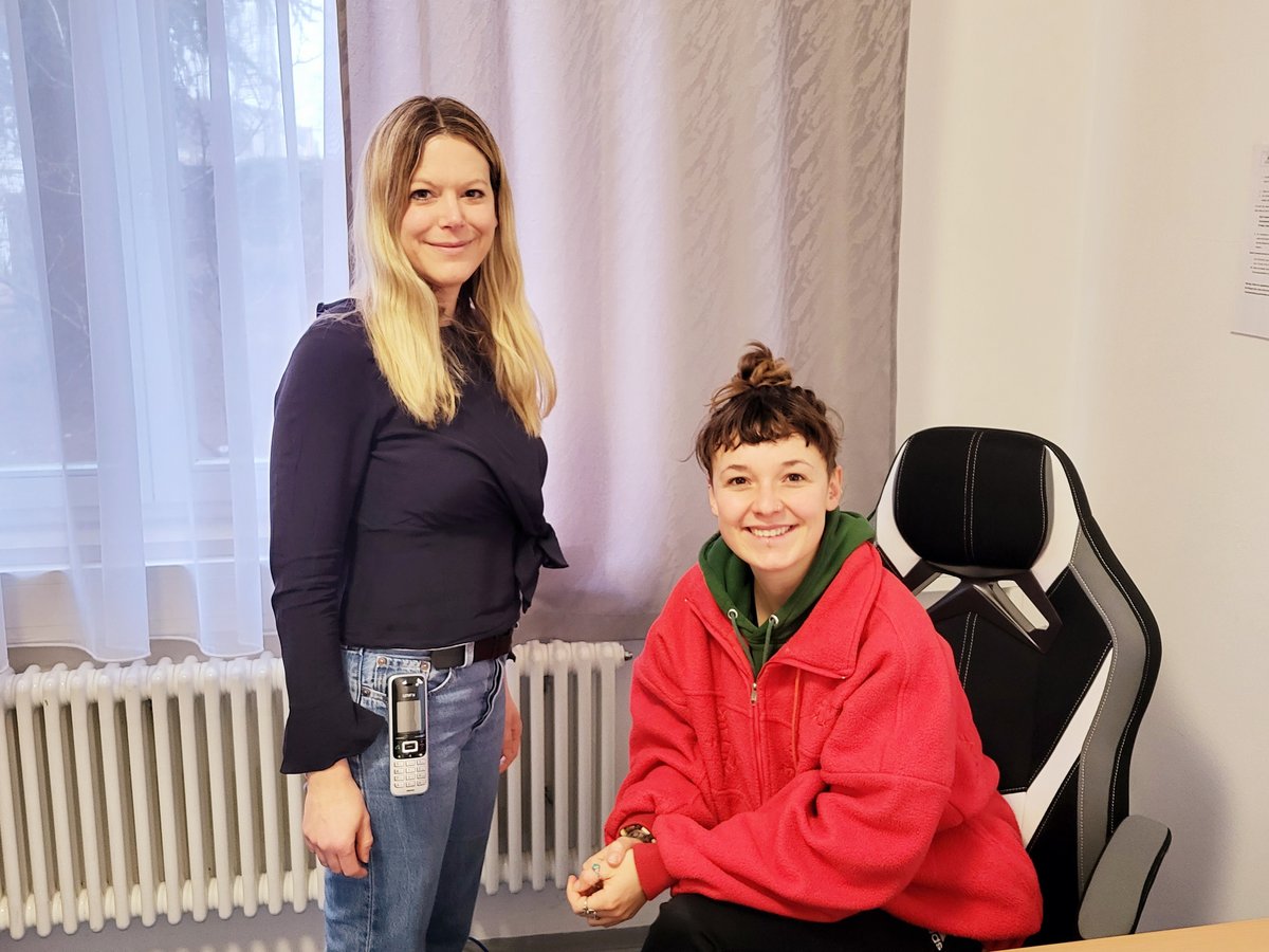 Zwei lächelnde Frauen im Büro. Eine blonde Frau steht, während eine andere Frau mit Dutt an einem Schreibtisch in roter Jacke sitzt.