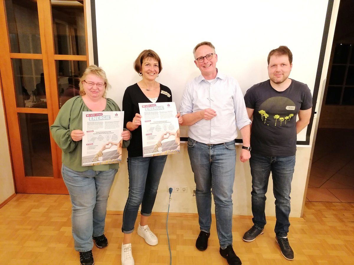 Vier lächelnde Personen, zwei Frauen und zwei Männer, halten Plakate mit der Aufschrift 'Wir teilen unsere ENERGIE'.