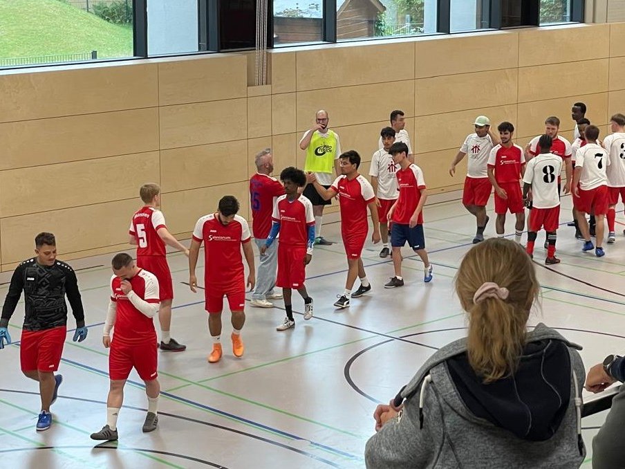 Hallenfußballspieler in roten und weißen Trikots versammeln sich auf einem Feld, während zwei Frauen im Vordergrund zusehen.