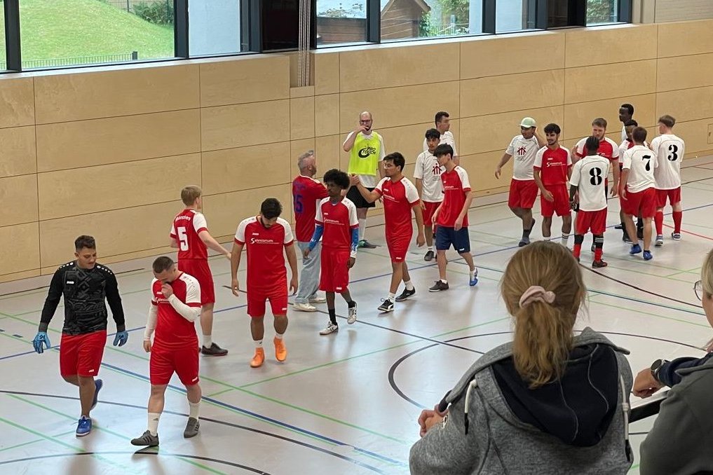 Hallenfußballspieler in roten und weißen Trikots versammeln sich auf einem Feld, während zwei Frauen im Vordergrund zusehen.