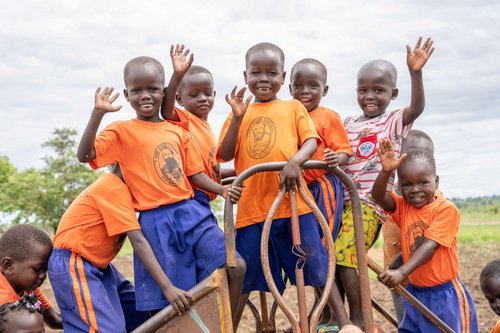 Fröhliche Kinder in orangefarbenen Schul-T-Shirts winken und lächeln auf einem Spielplatz.