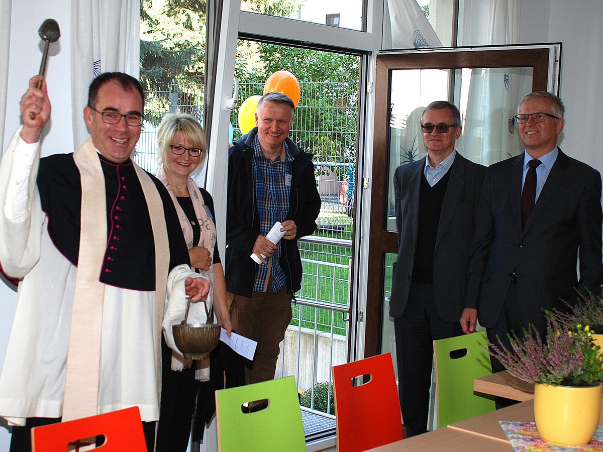 Ein lächelnder Priester mit Weihwassersprenger und -eimer, umgeben von lachenden Menschen bei einer Einweihungszeremonie.