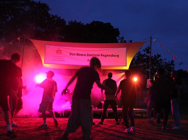 Menschen tanzen nachts im Freien unter roten und lila Bühnenlichtern. Im Hintergrund ein Banner des Don Bosco Zentrums Regensburg.