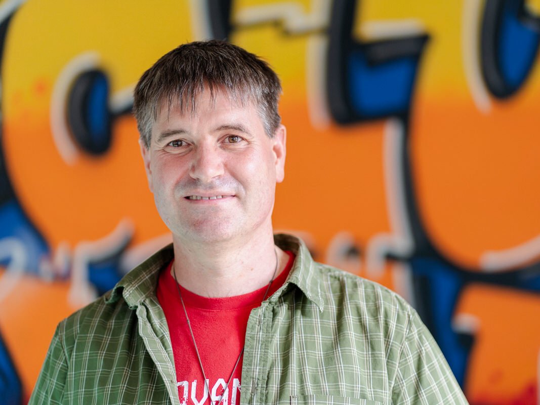 Ein lächelnder Mann mit kurzen grauen Haaren, einem grünen Karohemd über einem roten T-Shirt, vor einer bunten Graffitiwand.