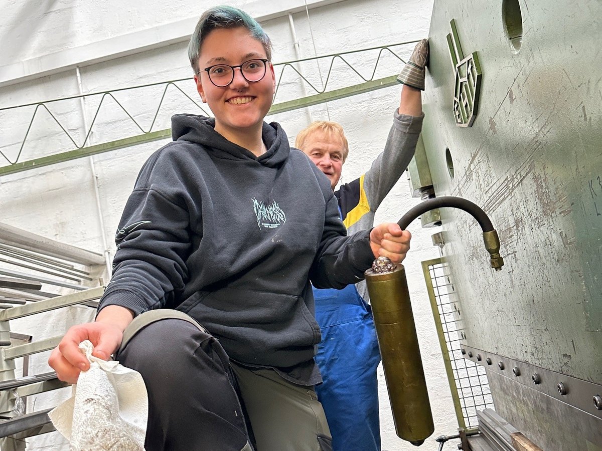 Eine junge Frau mit grauem Hoodie und Arbeitshose steht in einer Fabrikhalle und hält eine Gaskartusche in der Hand. Hinter steht ein Mann in Handwerkermontur. in einer Fabrikhalle 