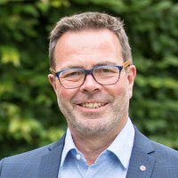 Porträt eines lächelnden Mannes mit Brille, hellblauem Hemd und blauem Sakko vor grünem Hintergrund.