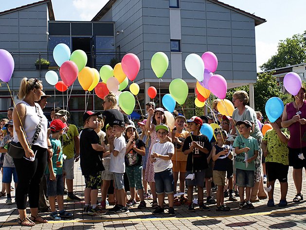 Kinder und Erwachsene stehen draußen und halten viele bunte Ballons bei einem sonnigen Fest.