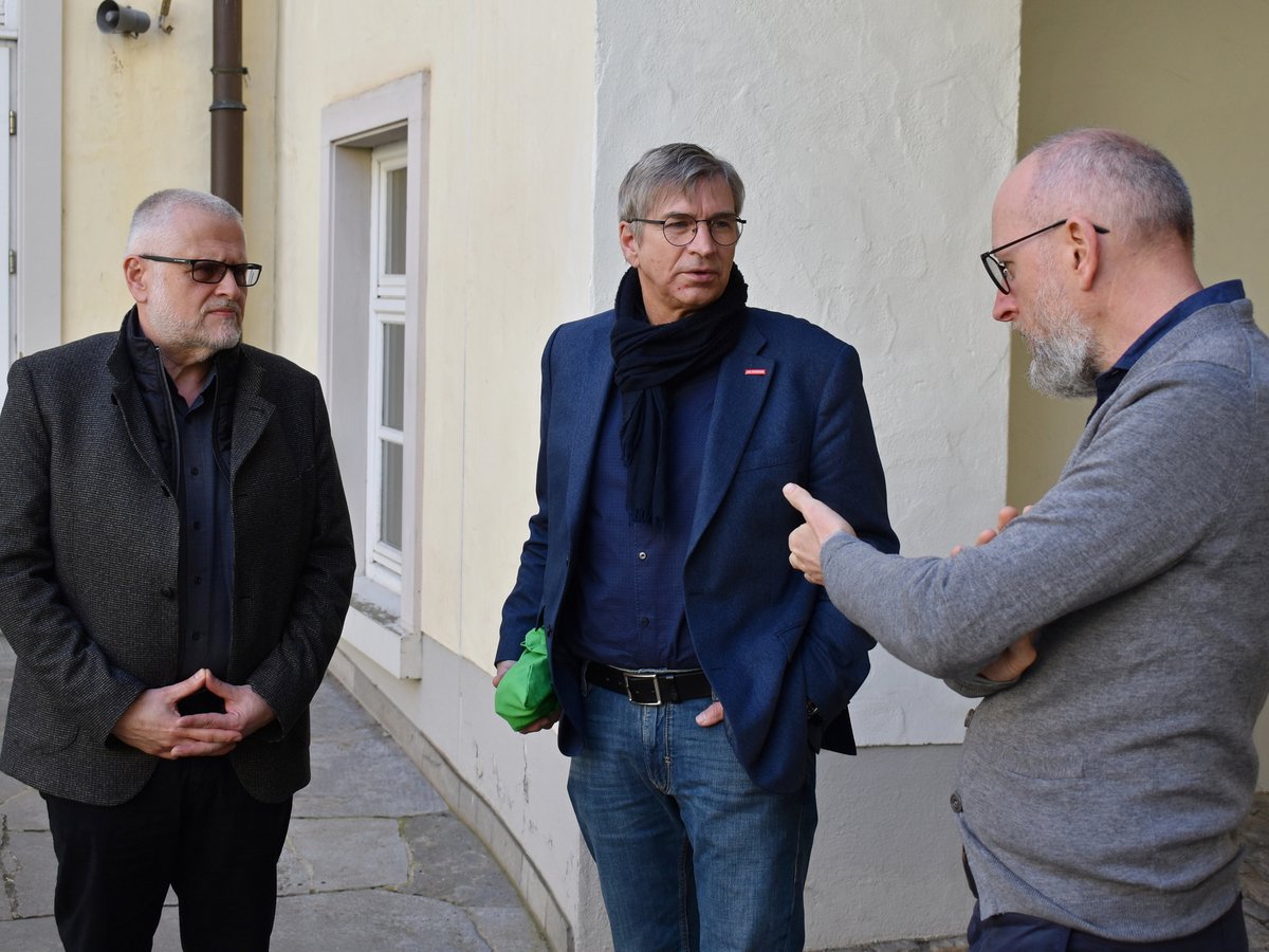 Drei Männer unterhalten sich im Freien vor einem hellen Gebäude. Einer gestikuliert, die anderen hören zu.