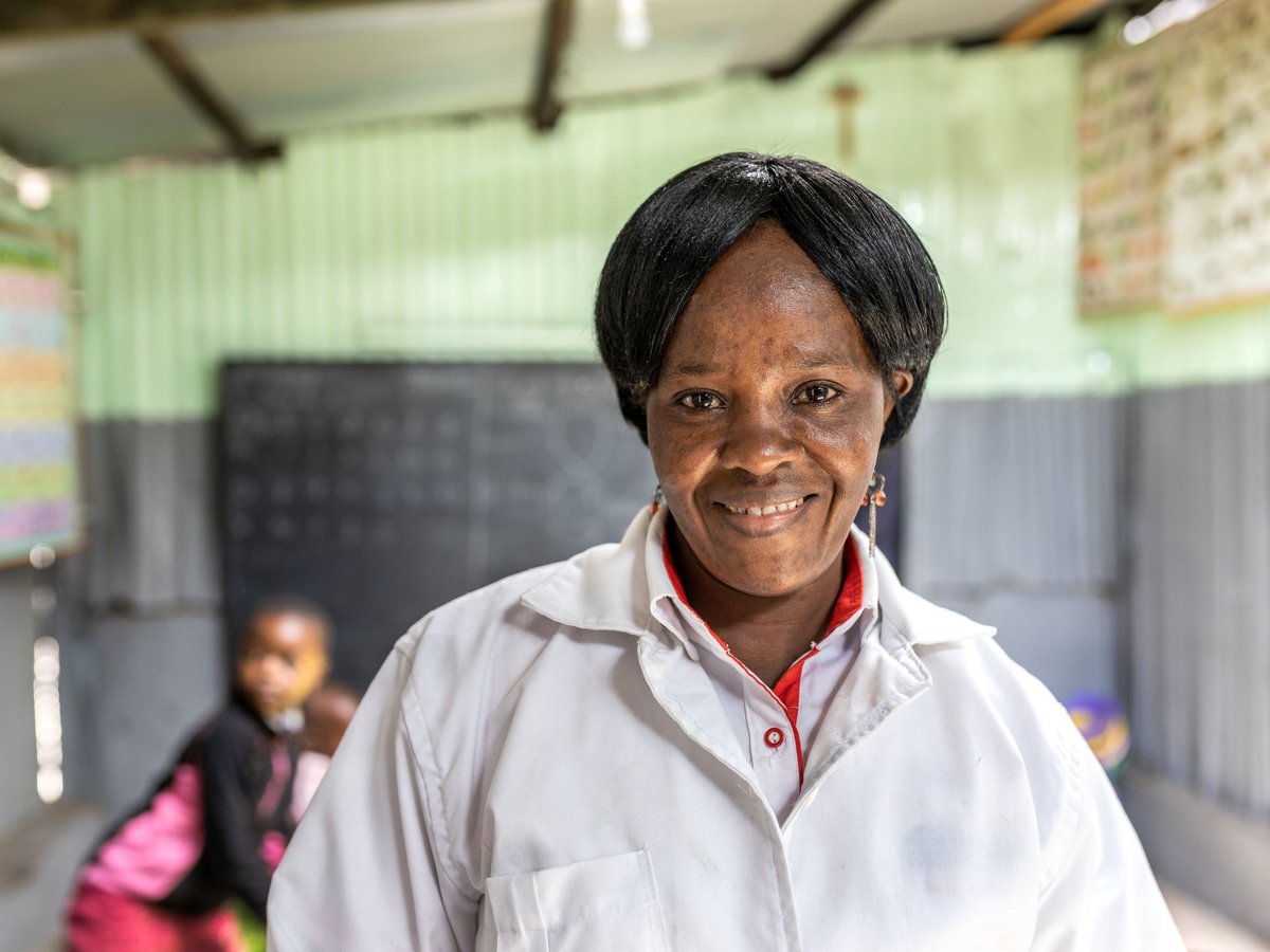Nahaufnahme einer lächelnden Schwarzen Frau im weißen Kittel in einem Klassenzimmer mit einem unscharfen Kind im Hintergrund.