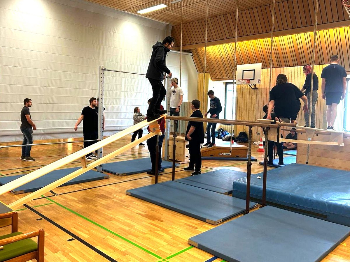 Jugendliche trainieren in einer Turnhalle an einem Parkour-Parcours mit Barren, Schwebebalken und Sprungkästen, umgeben von Weichbodenmatten.