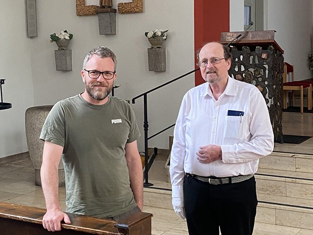 Zwei Männer, ein jüngerer mit Bart und ein älterer, stehen in einem Kirchenraum mit religiöser Ausstattung.