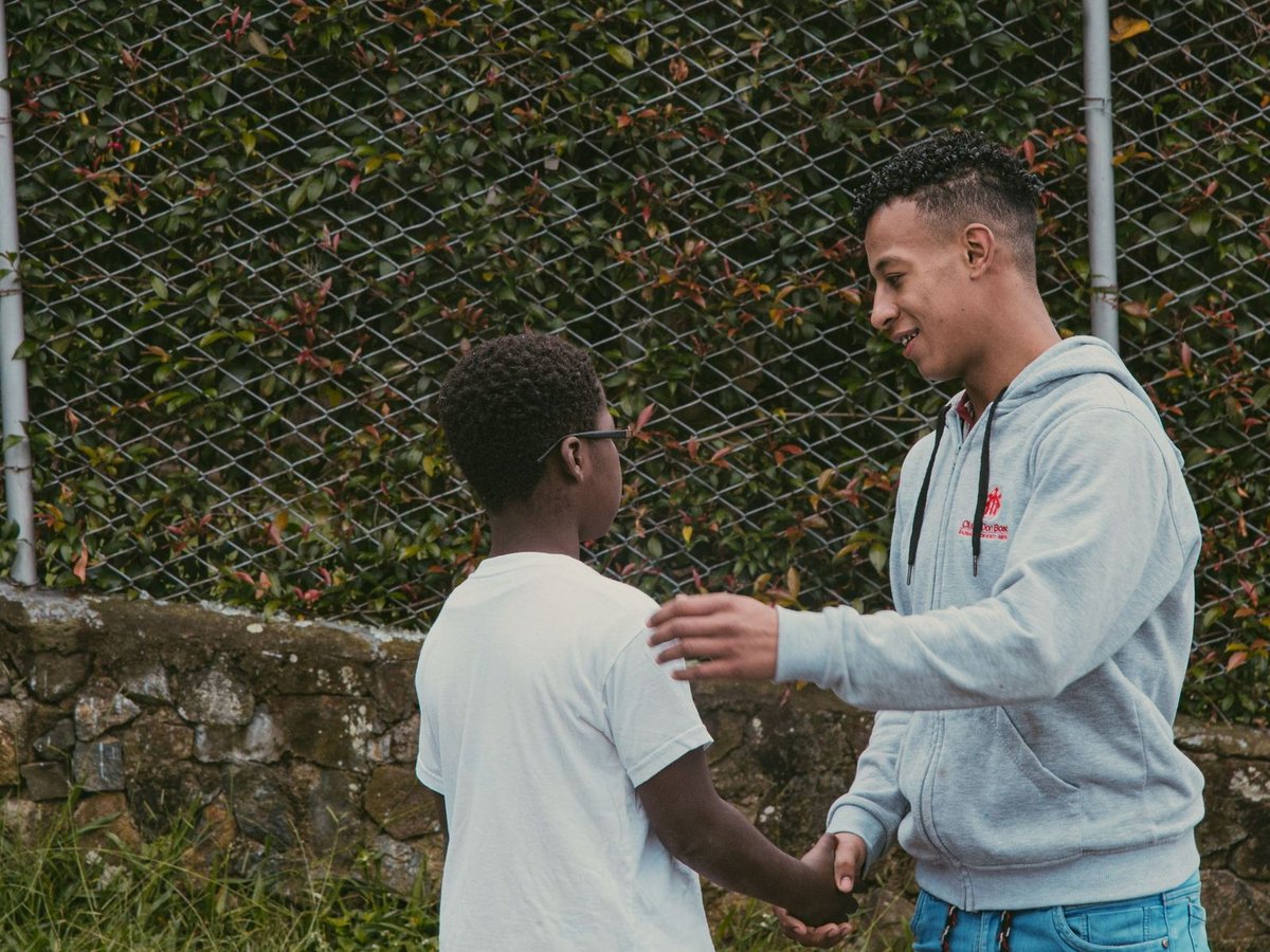 Zwei Jungen, ein älterer und ein jüngerer, schütteln sich freundlich die Hände und teilen einen unterstützenden Moment im Freien vor einem bewachsenen Zaun.