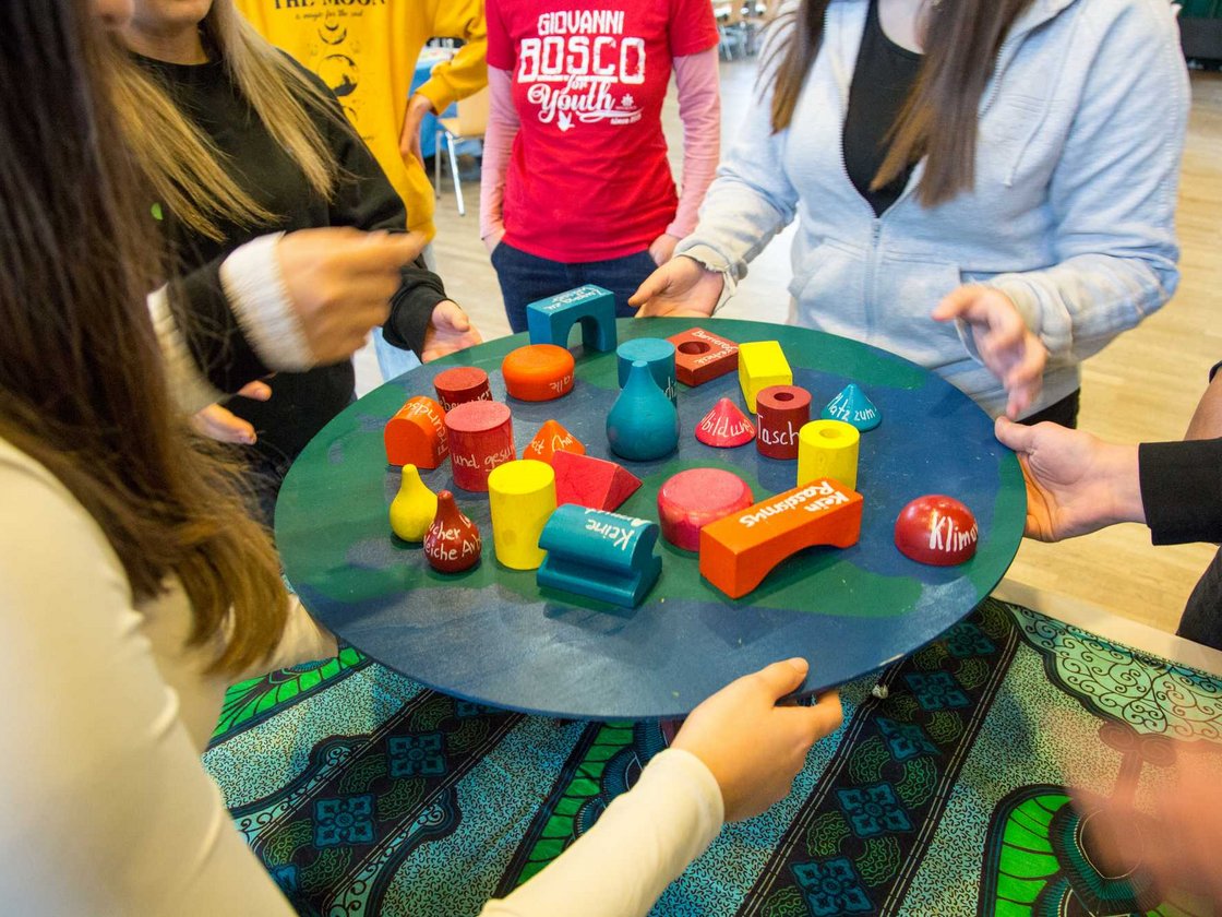 Jugendliche halten gemeinsam eine Erdkugel-Platte mit bunten Blöcken, die soziale Themen wie Klima und Rassismus darstellen.