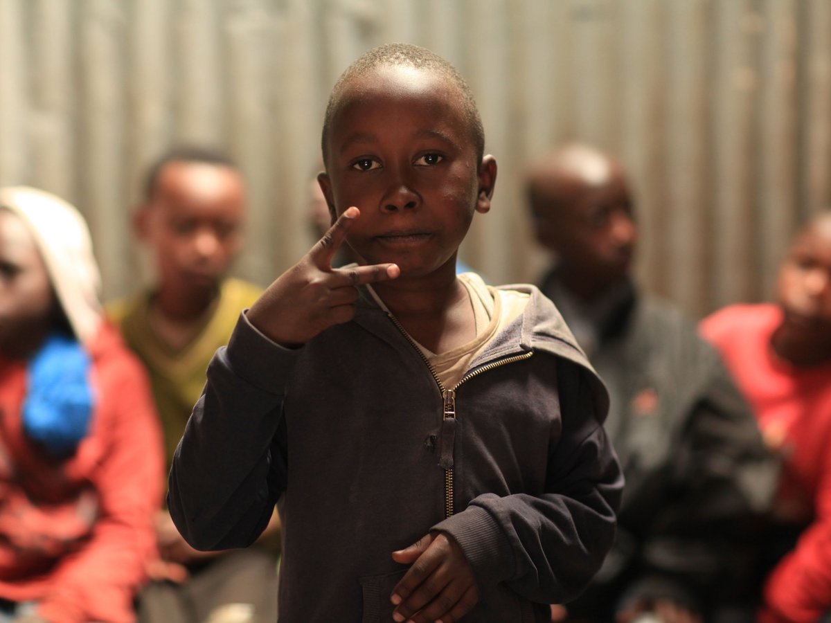 Ein dunkelhautiges Kind in Kapuzenpullover zeigt das Friedenszeichen und blickt in die Kamera. Unscharfe Personen im Hintergrund.