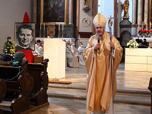 Ein Bischof in goldener liturgischer Kleidung spricht mit Mikrofon und Krummstab in einer Kirche vor Gemeindemitgliedern und einem Porträt.