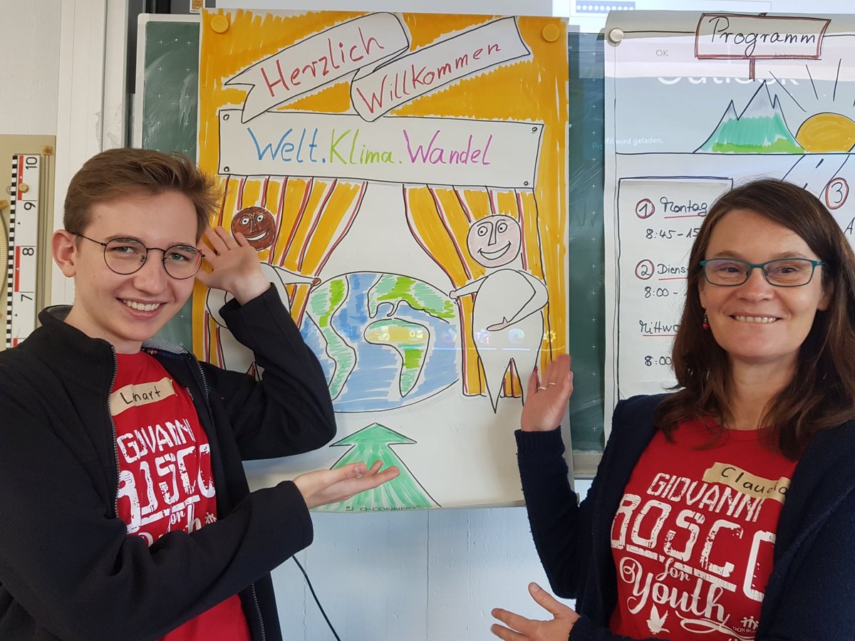 Lächelnder Mann und Frau in 'Giovanni Bosco for Youth'-Shirts zeigen auf ein 'Willkommen Welt.Klima.Wandel'-Plakat.