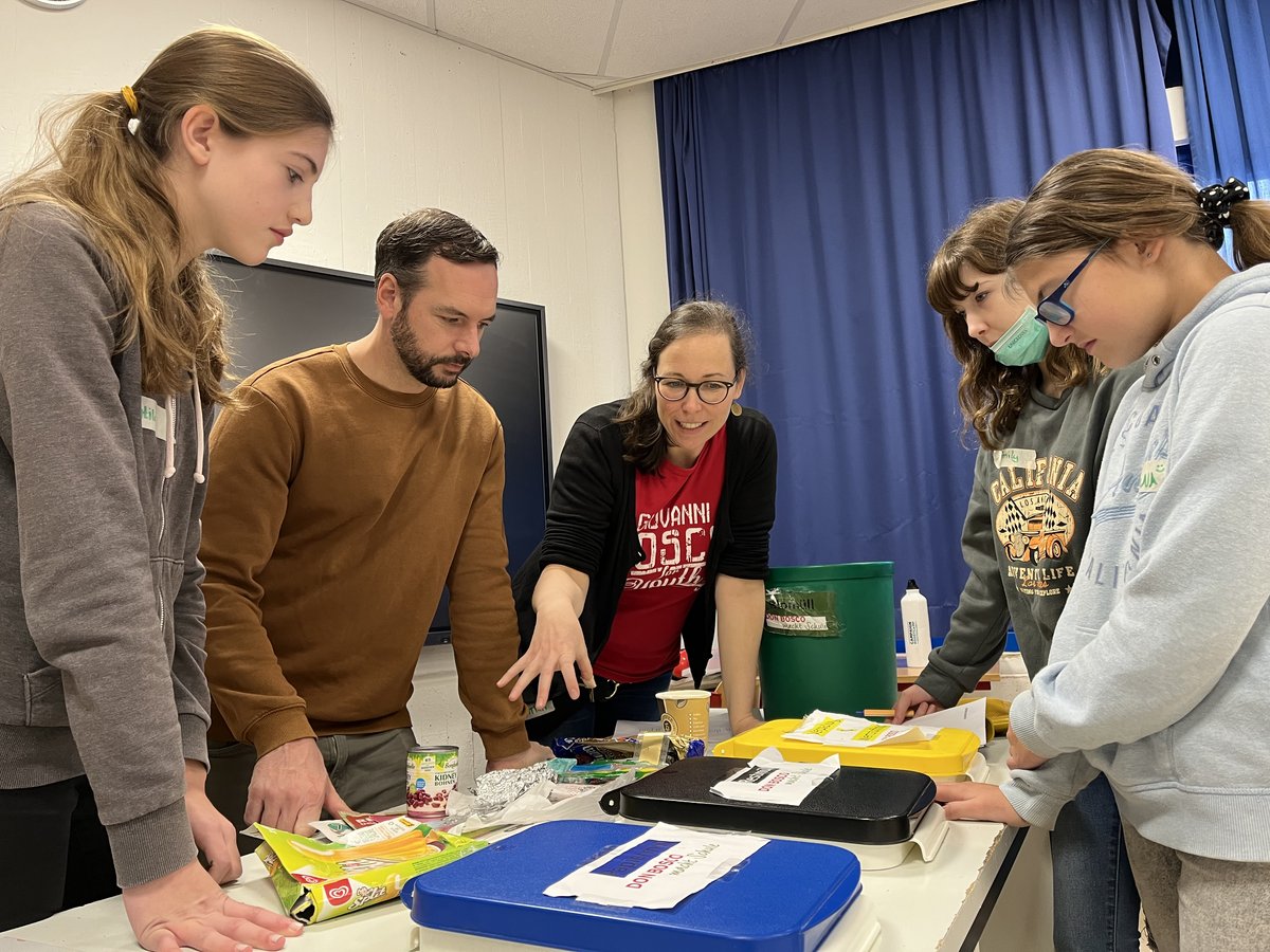 Erwachsene und Schüler versammeln sich um einen Tisch mit Lebensmitteln und Behältern, diskutieren und lernen.