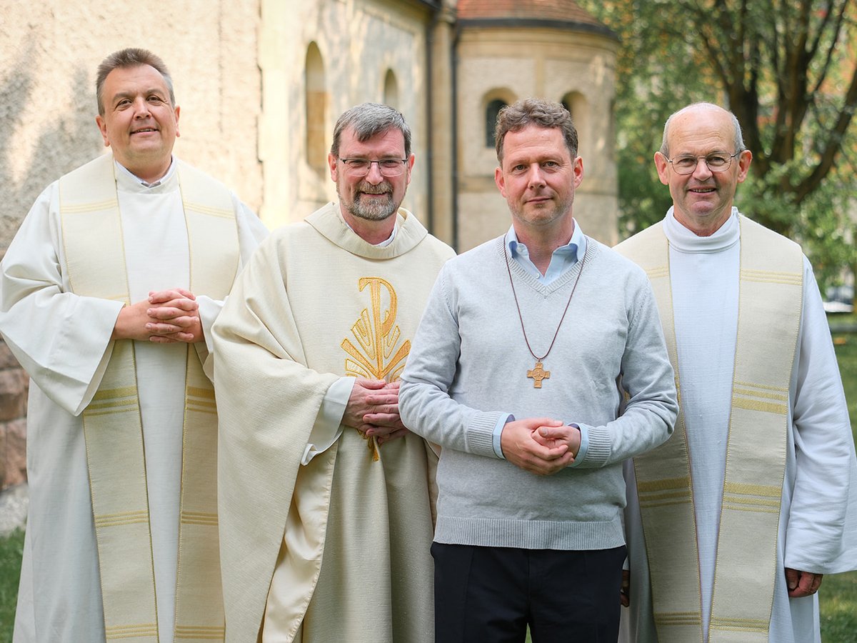Vier Männer, drei in Priestergewändern und einer mit Kreuzkette, stehen lächelnd vor einem Kirchengebäude.