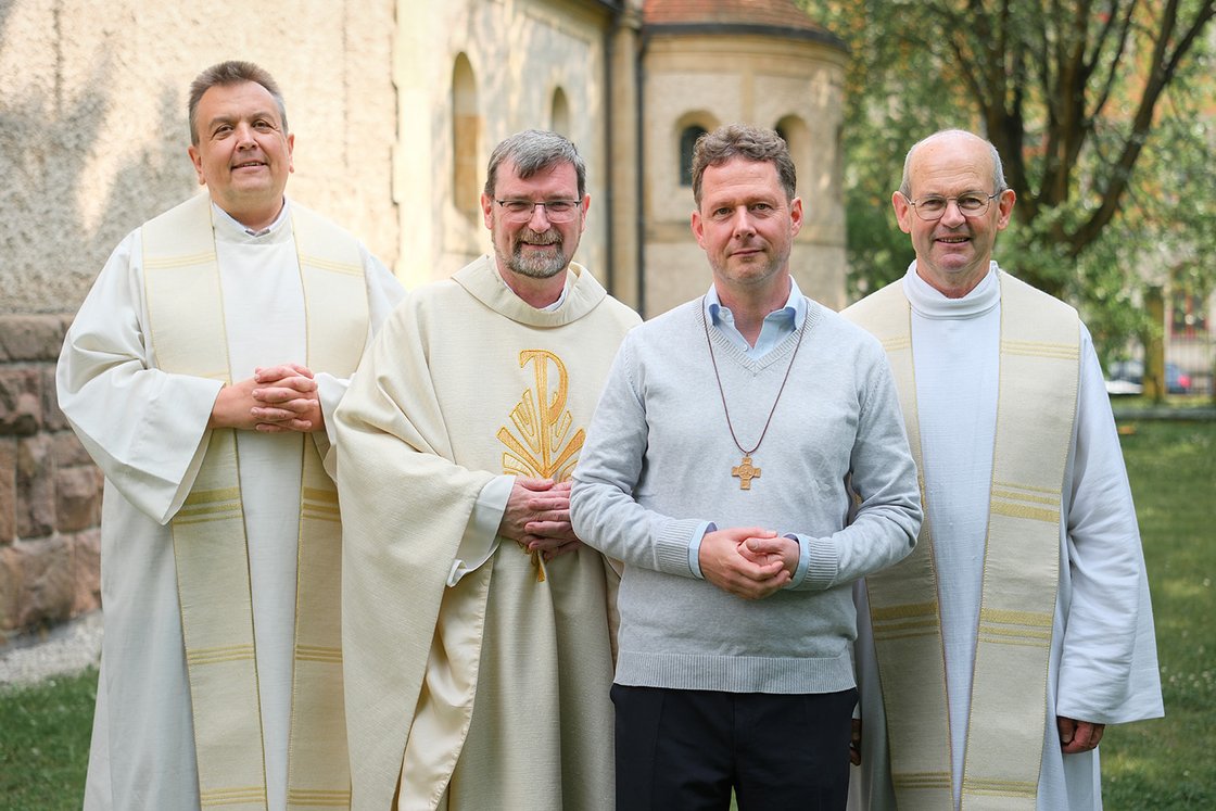 Vier Männer, drei in Priestergewändern und einer mit Kreuzkette, stehen lächelnd vor einem Kirchengebäude.