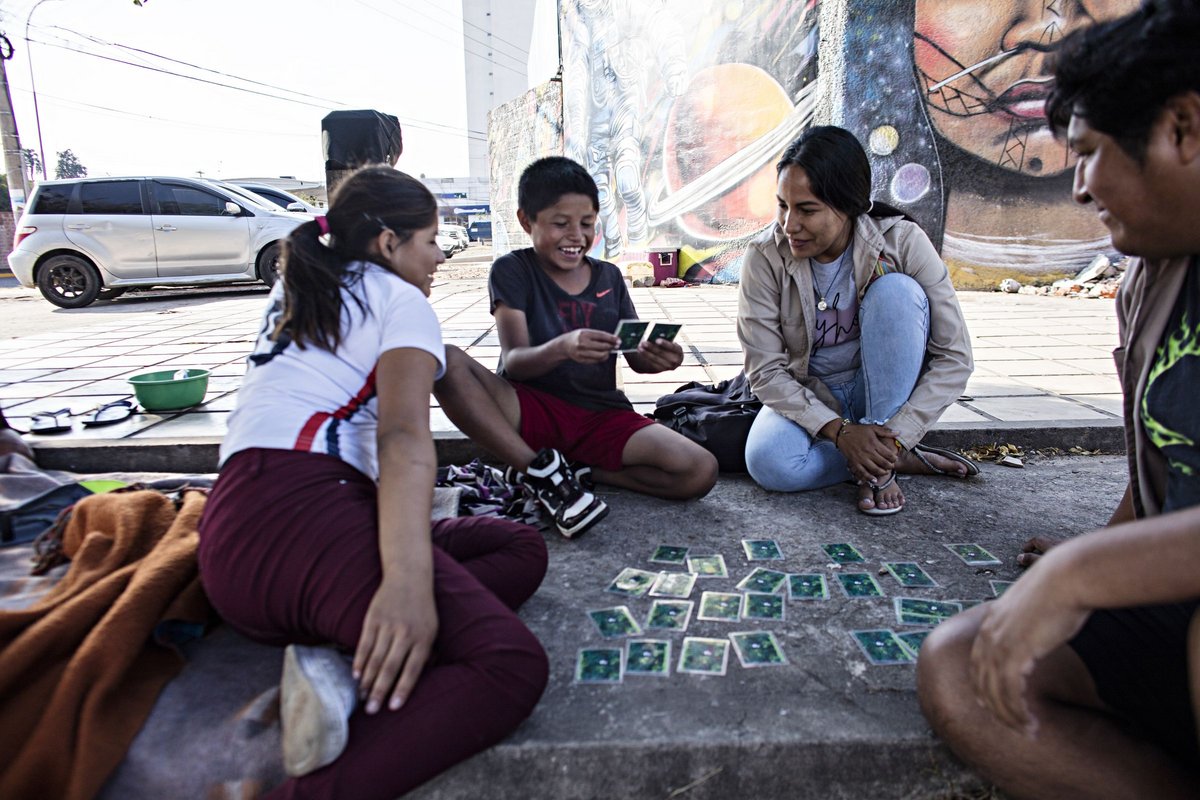 Vier lachende Menschen spielen Karten auf dem Bürgersteig vor einem farbenfrohen Wandgemälde, der Junge in der Mitte hält Karten.