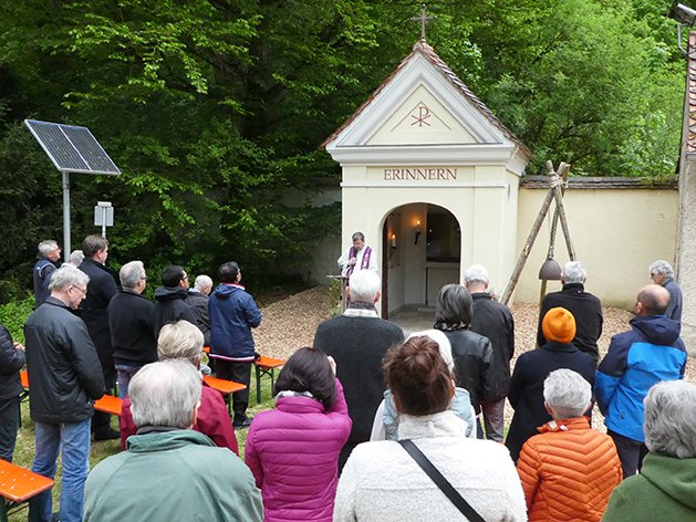 Menschen versammeln sich vor einer Kapelle mit der Aufschrift „Erinnern“, wo ein Priester eine Andacht hält.