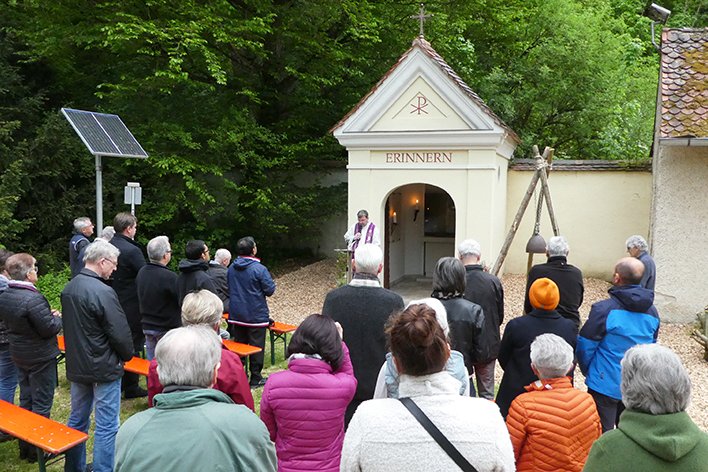 Menschen versammeln sich vor einer Kapelle mit der Aufschrift „Erinnern“, wo ein Priester eine Andacht hält.
