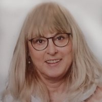 Eine lächelnde blonde Frau mit Brille und einem mehrfarbigen Kapuzenpullover vor hellem Hintergrund.