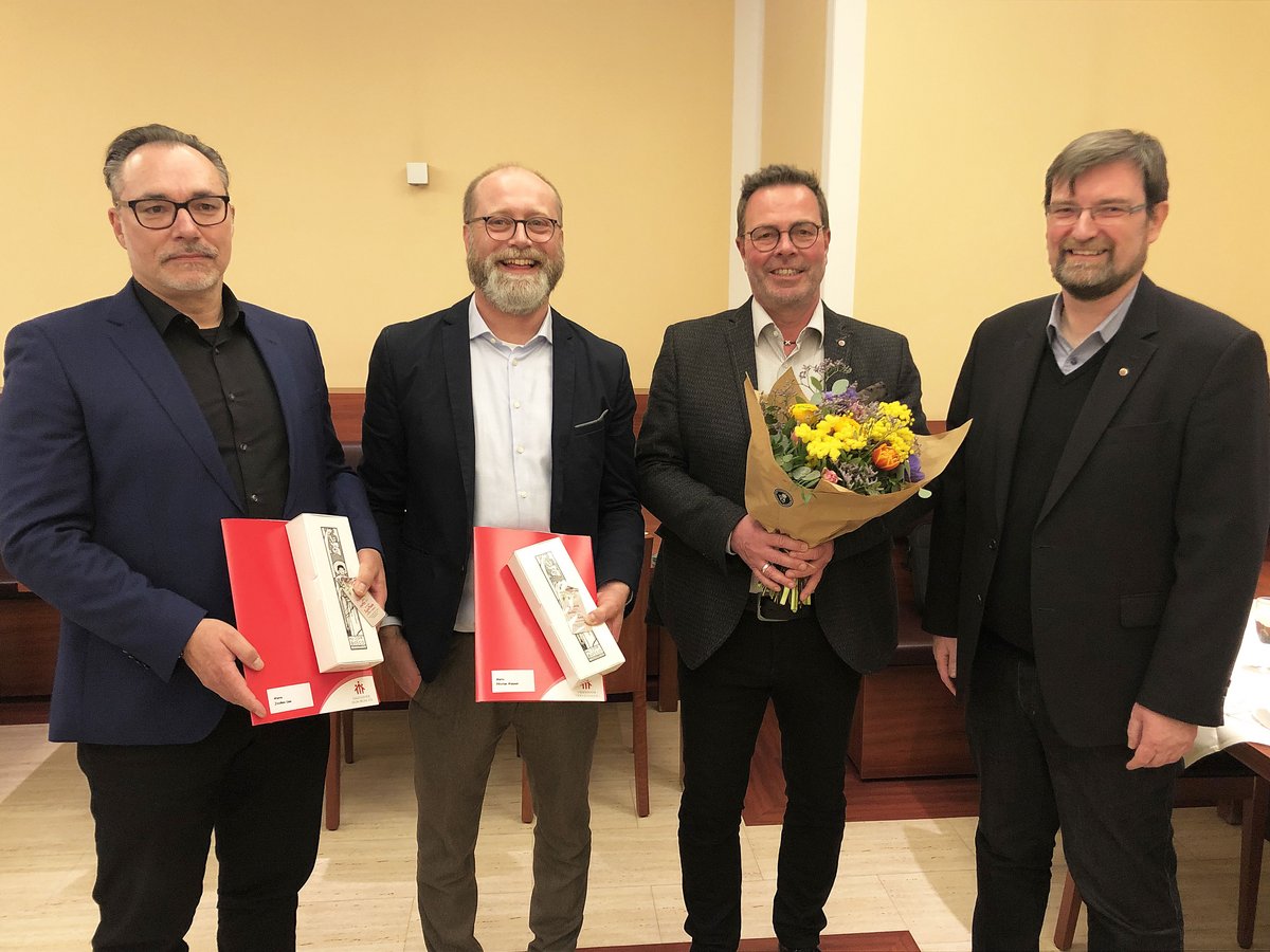 Vier lächelnde Männer bei einer Veranstaltung. Zwei halten rote Mappen und Geschenkboxen, einer einen Blumenstrauß.