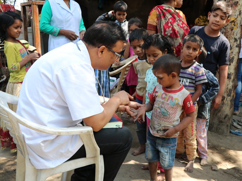 Ein Arzt im weißen Hemd und mit Stethoskop untersucht die Hand eines Kindes. Viele Kinder schauen im Freien zu.