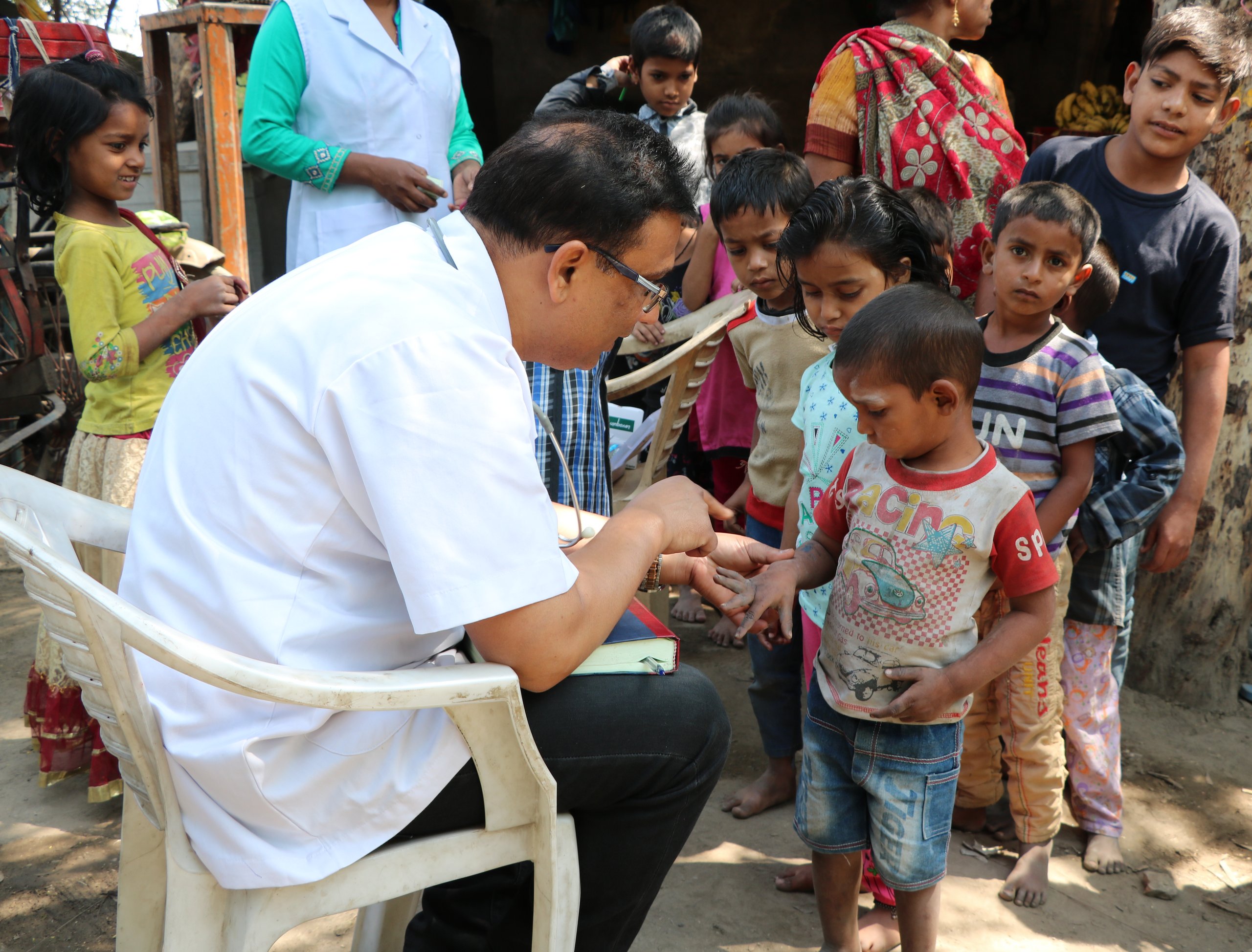 Ein Arzt im weißen Hemd und mit Stethoskop untersucht die Hand eines Kindes. Viele Kinder schauen im Freien zu.