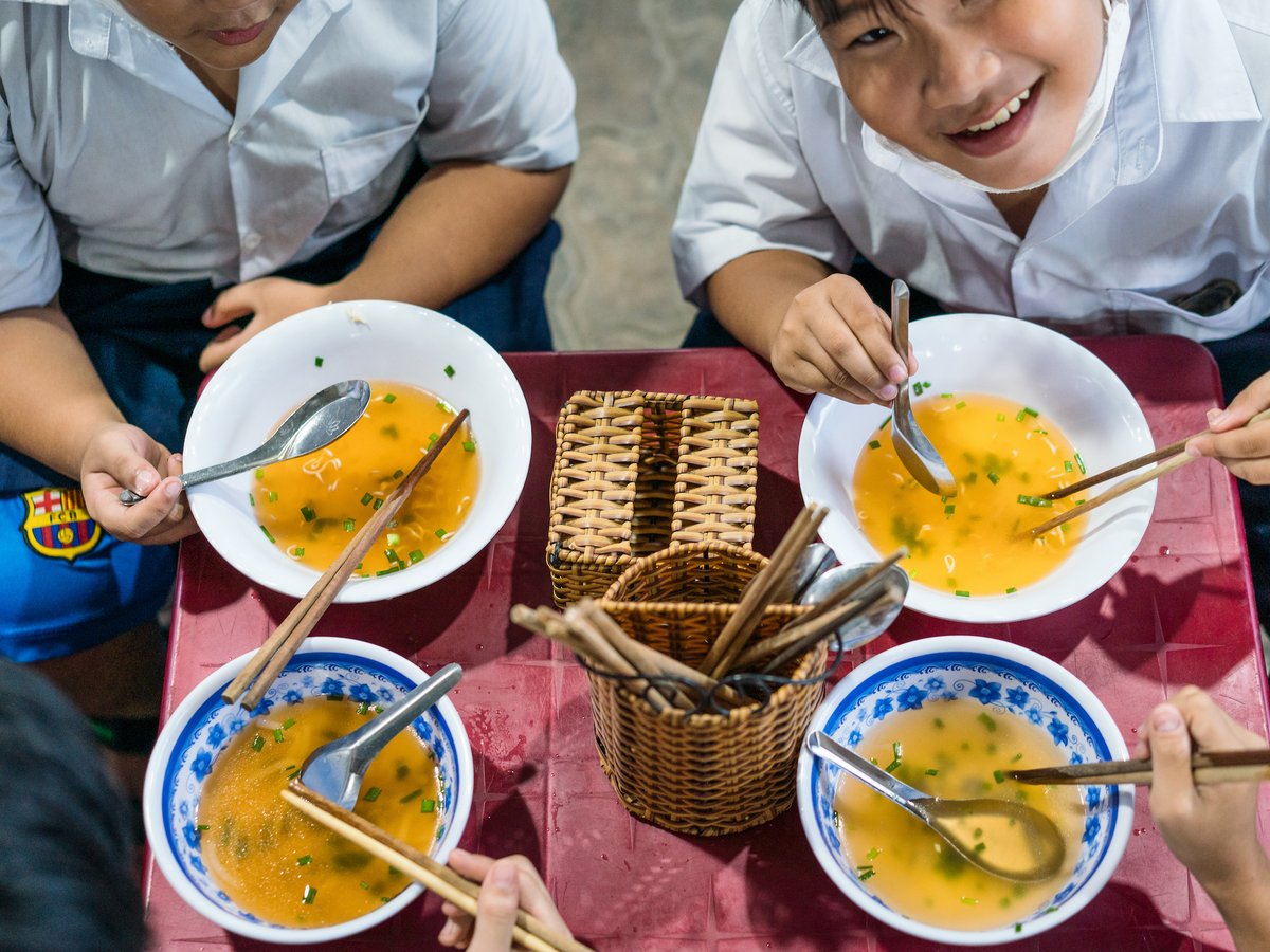 Lächelnde Kinder in Schuluniformen essen Suppe mit Stäbchen und Löffeln an einem roten Tisch.