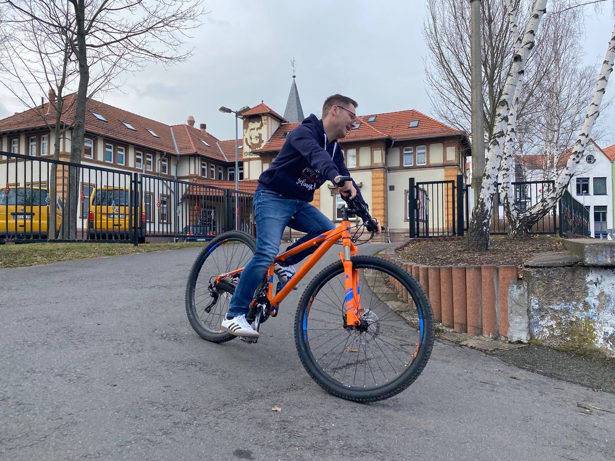 Lächelnder Mann in Hoodie und Jeans fährt orangefarbenes Mountainbike auf Weg vor historischen Gebäuden und Bäumen.