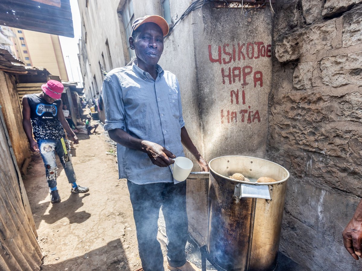 Mann mit oranger Mütze verkauft Essen aus dampfendem Topf auf Straße mit Graffiti. Ein weiterer Mann im Hintergrund.