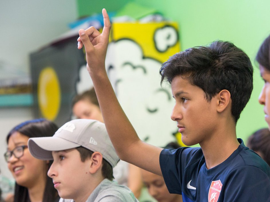 Ein junger Junge in einem blauen Hemd hebt seine Hand in einem Klassenzimmer, bereit, eine Frage zu beantworten.