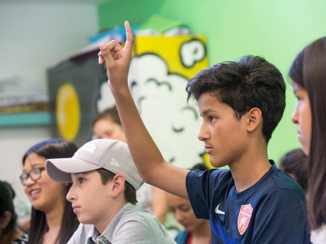 Ein junger Junge in einem blauen Hemd hebt seine Hand in einem Klassenzimmer, bereit, eine Frage zu beantworten.
