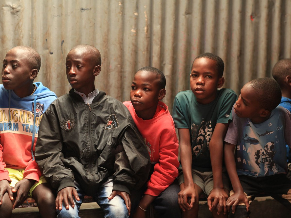 Eine Gruppe von sechs jungen afrikanischen Jungen sitzt ernst blickend eng zusammen vor einer Wellblechwand.