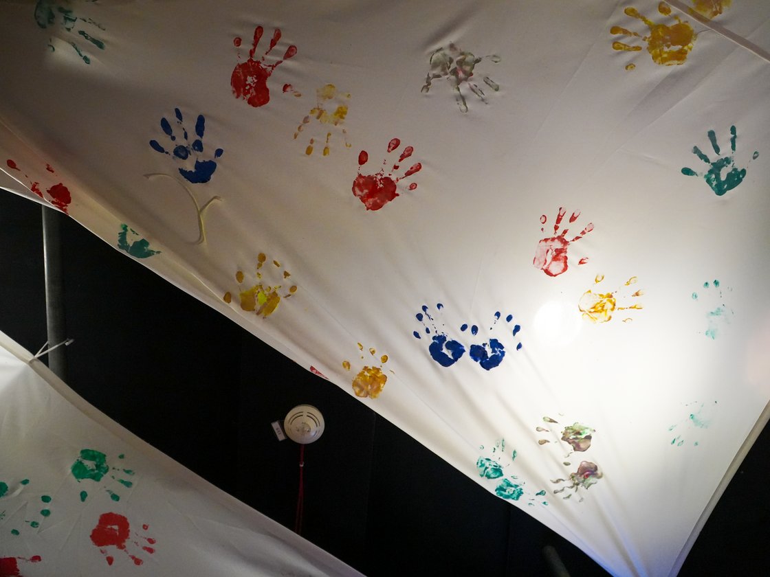 Weiße Stoffdecke bedeckt mit bunten Handabdrücken (Rot, Blau, Gelb, Grün), von einem Licht beleuchtet.