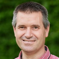 Ein lächelnder Mann mittleren Alters mit kurzen, grau melierten Haaren und einem rot karierten Hemd vor grünem Hintergrund.