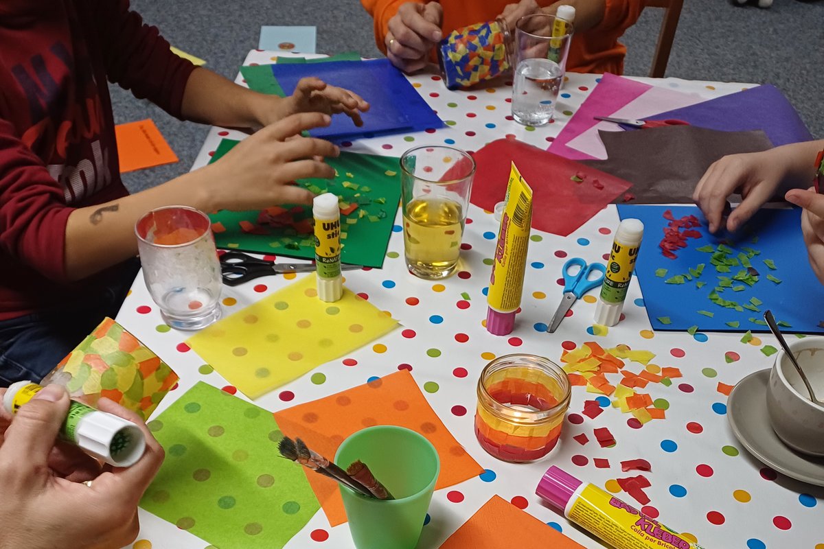 Kinder sitzen um einen Tisch und basteln Erinnerungslichter aus Gläsern und buntem Papier