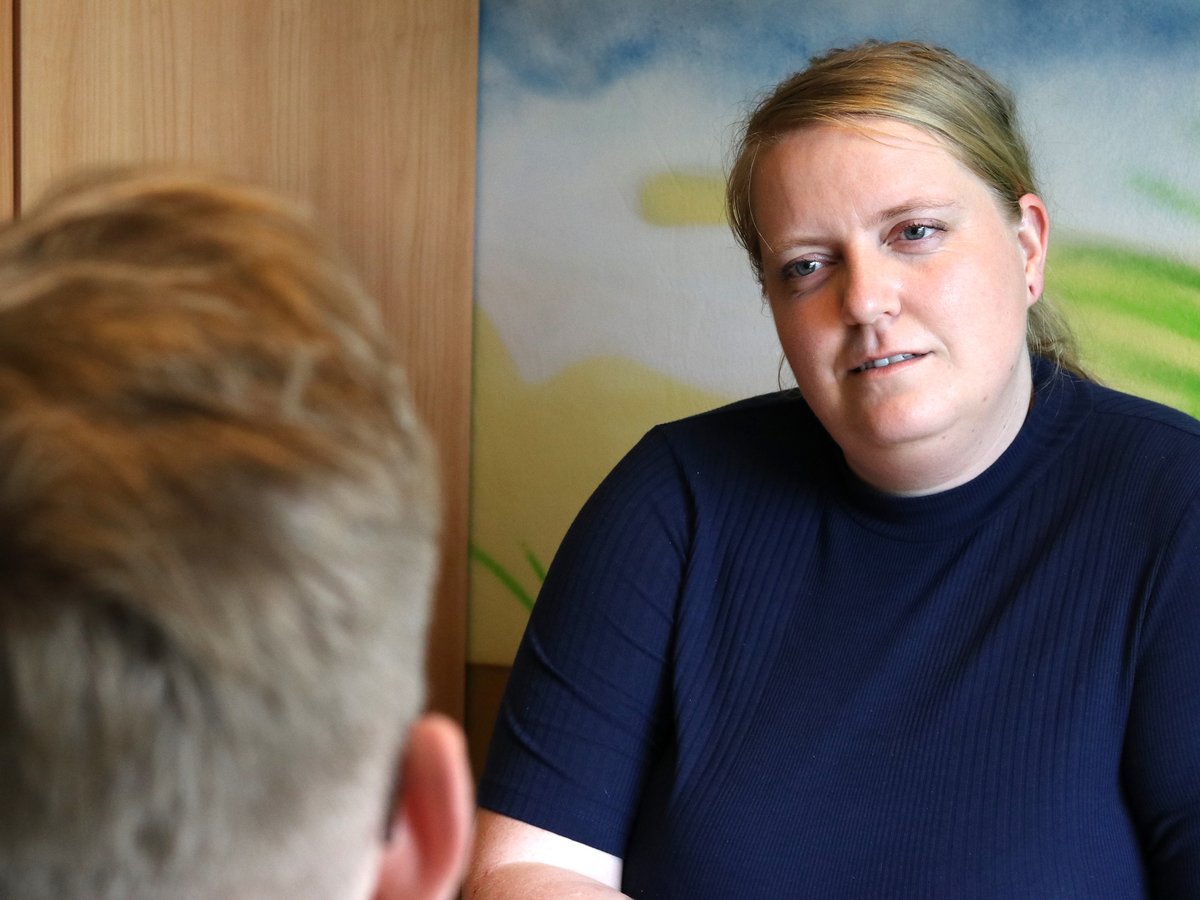 Eine blonde Frau in einem dunkelblauen gerippten Oberteil lächelt, blickt an der unscharfen Person im Vordergrund vorbei.