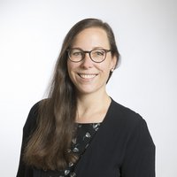 Lächelnde Frau mit langen braunen Haaren und Brille, trägt ein dunkles Oberteil, vor weißem Hintergrund.