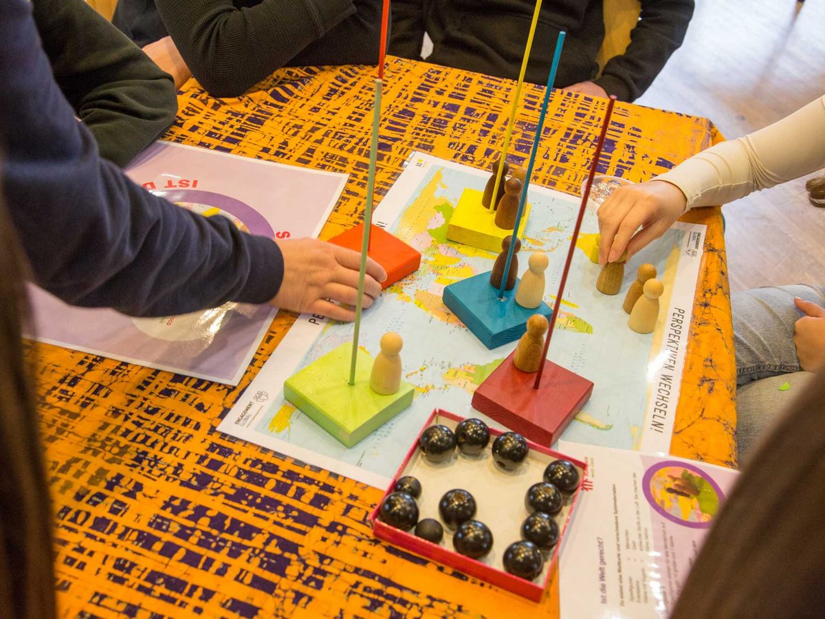 Menschen spielen ein Brettspiel auf einer Weltkarte mit Holzfiguren, farbigen Blöcken und Stäben, um globale Perspektiven zu erkunden.